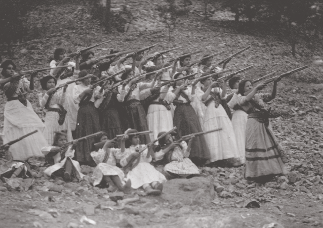 Black and white photo of a group made up of about 20 women and girls dressed in traditional Mexican dress and armed with rifles. 