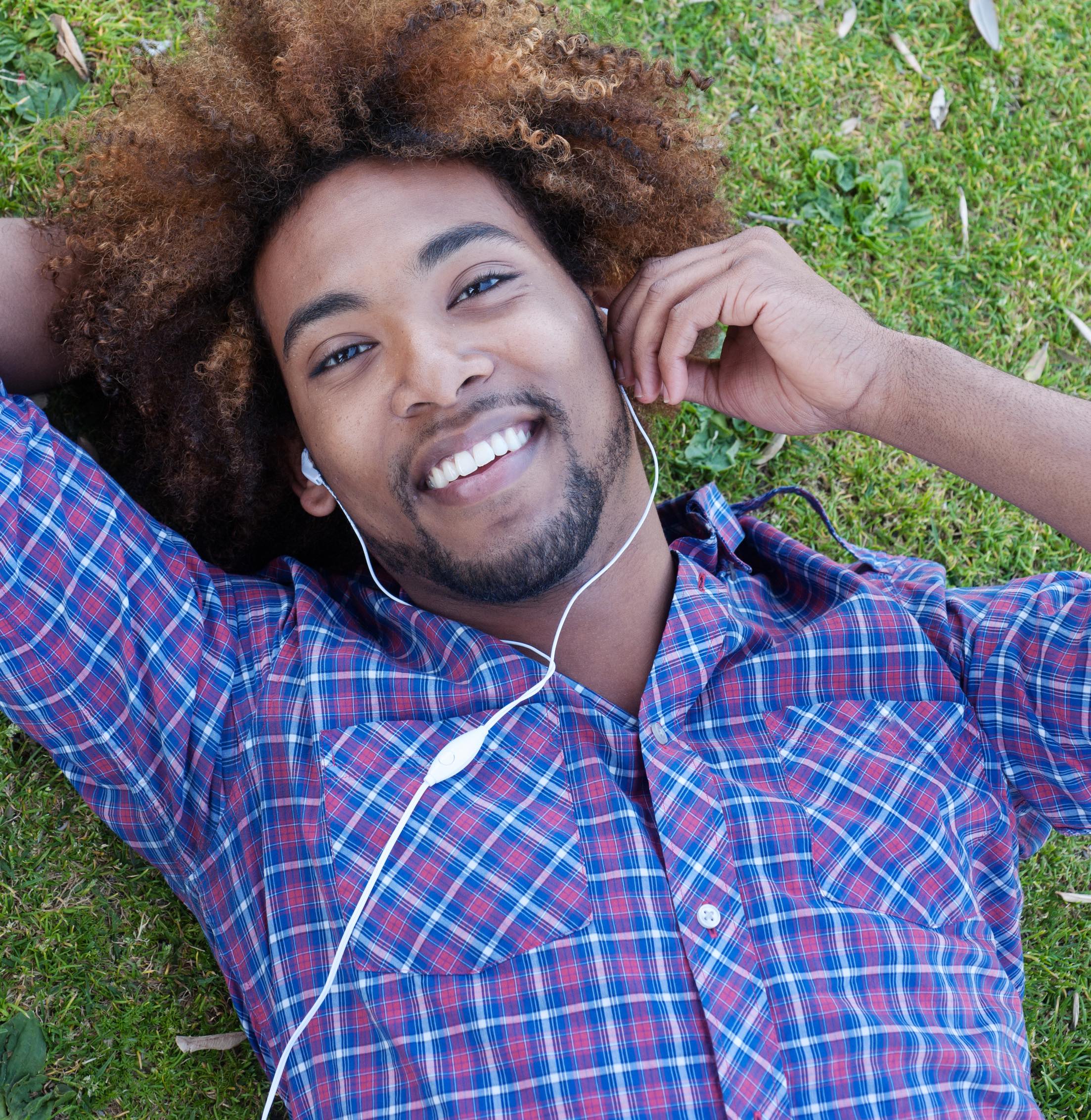 A smiling man listening to music with earphones while lying in the grass.