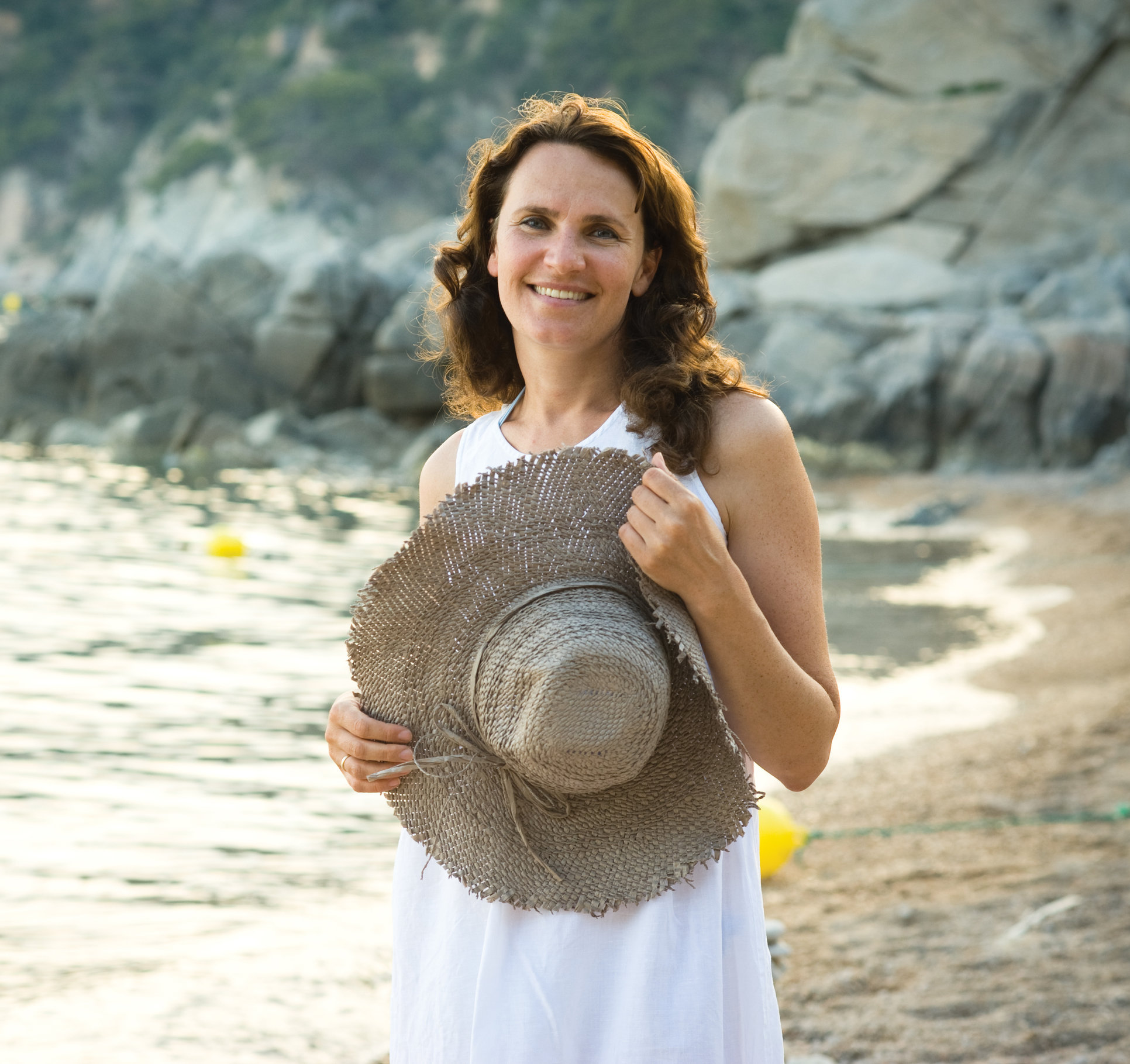 A smiling woman posing for a photo on the beach. She wears a white dress and holds a straw hat.