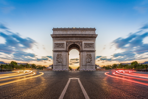 Arc de Triomphe à Paris, illuminé au crépuscule, entouré de voitures en mouvement et de nuages.