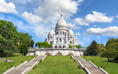 Basilique du Sacré-Cœur à Paris, entourée de jardins et d'escaliers, sous un ciel bleu avec quelques nuages.