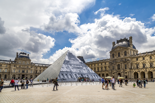 La pyramide du Louvre entourée de visiteurs, avec le musée historique en arrière-plan sous un ciel nuageux.