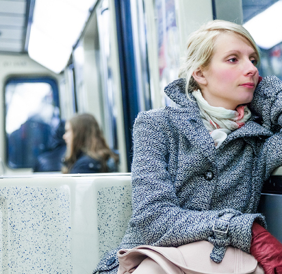 A woman with a blank expression riding a train. She leans on her arm against the window of the train. The woman's eyes are half-open. 