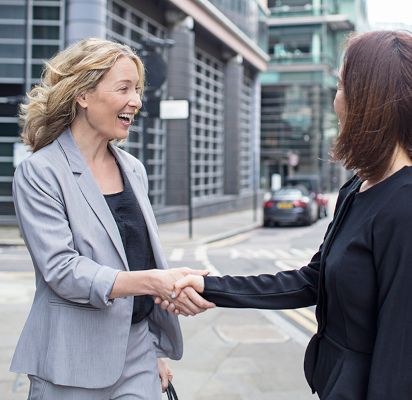 Two women shaking hands. One woman raises her eyebrows as she smiles happily at the other woman.